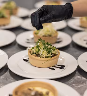 En kock som bär en svart handske garnerar en rad små salta tårtor. Tårtorna är fyllda med grönt och andra ingredienser, placerade på vita tallrikar med en klick mörk sås, arrangerade på en yta av rostfritt stål.