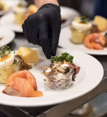 Närbild av en kocks hand som bär en svart handske och garnerar tallrikar med färska örter. Tallrikarna innehåller rökt lax, ett kokt ägg, blandade grönsaker och en burk svampsallad, placerad på en yta av rostfritt stål.