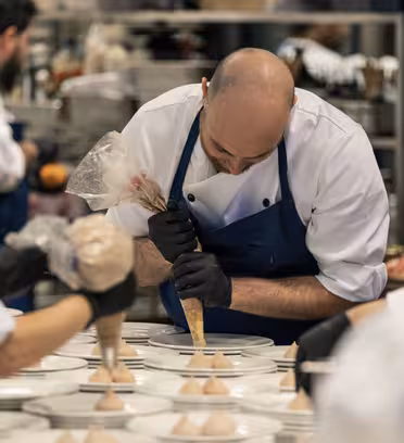 En kock i förkläde sprider choklad på en tallrik i ett hektiskt kök, omgiven av andra kockar. Borden håller förberedda desserter redo för plätering. Scenen är full av kulinariska aktiviteter.