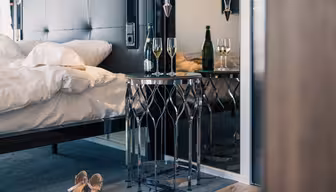 Hotellrum med en prydligt bäddad säng och ett litet, runt glasbord. På bordet står två glas och en flaska mousserande vin. Ett par snygga högklackade skor placeras på golvet bredvid en plyschmatta.