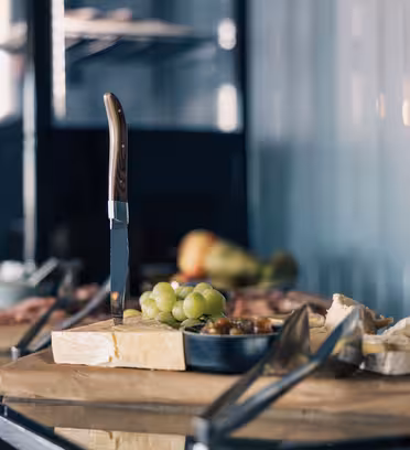 En köksbänk med en mängd olika ostar, en kniv som står upprätt på en skärbräda och en liten skål med gröna druvor. Bakgrunden är suddig, vilket framhäver sortimentet av mat i förgrunden.