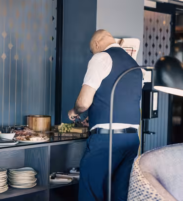 En man i vit skjorta och blå väst står vid ett buffébord på en restaurang. Han väljer mat inklusive vindruvor och skivat kött. Hyllor under bordet rymmer tallrikar. Miljön har en modern, svagt upplyst interiör med elegant inredning.