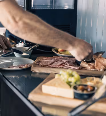 En person förbereder en tallrik med diverse kött på en självbetjäningsstation. Disken innehåller skivat kött, ost och ett litet fat med pickles. Tallrikar är staplade i närheten, och inställningen verkar vara en buffé eller delikatessdisk.
