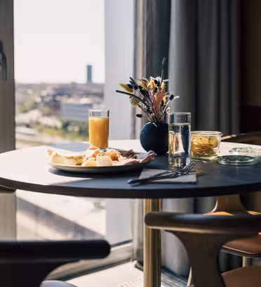 Ett frukostbord vid ett fönster med utsikt över staden. På bordet finns en tallrik med diverse mat, ett glas apelsinjuice, ett glas vatten, en liten burk med frukt och en vas med torkade blommor. Trästolar omger bordet.