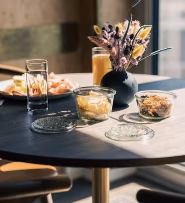 Runt bord med fat i solljus, med ett glas vatten, apelsinjuice, bröd på en tallrik och burkar med mat. En liten vas med dekorativa blommor står i mitten. Moderna stolar och en fönstervy i bakgrunden.