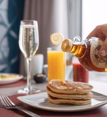 En hand häller sirap på en bunt pannkakor på en vit tallrik. Det finns ett glas apelsinjuice och en flöjt mousserande vin i närheten. En kopp kaffe och en tallrik mat sitter i bakgrunden på ett bord med en röd bordstablett.