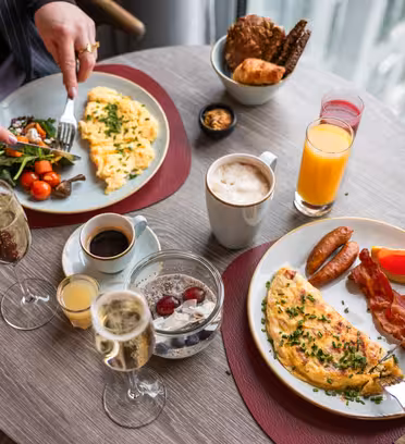Två personer äter frukost vid ett runt bord. Tallrikarna innehåller omeletter, körsbärstomater, korv, bacon och rostat bröd. Drycker består av apelsinjuice, kaffe och champagne. En person lägger peppar till en omelett. Inställningen är vid ett fönster.
