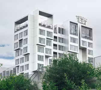 Modern flervåningsbyggnad med unik kubisk design och stora fönster, mot en molnig himmel. Frodiga gröna träd och ett metallstängsel syns i förgrunden.