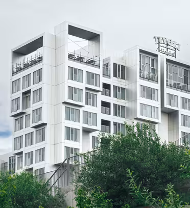 Modern flervåningsbyggnad med unik kubisk design och stora fönster, mot en molnig himmel. Frodiga gröna träd och ett metallstängsel syns i förgrunden.