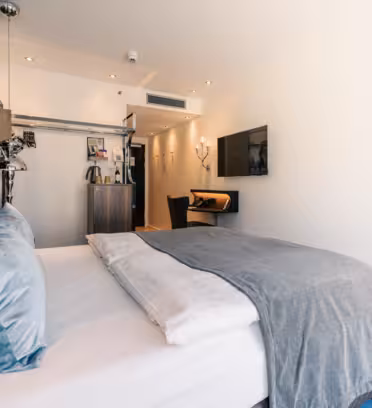 Ett modernt hotellrum med en snyggt bäddad säng med vita sängkläder och grå och blå kuddar. En grå filt är draperad över sängen. I rummet finns en väggmonterad TV, ett litet skrivbord, stol och en kaffestation med hylla. Belysningen är varm och inbjudande.