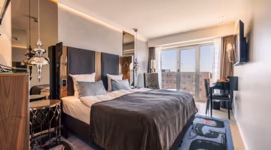 Modernt hotellrum med dubbelsäng, grå sängkläder och vadderade sänggavel. Stora fönster erbjuder utsikt över staden. Ett skrivbord med en stol finns bredvid fönstret och en väggmonterad TV vetter mot sängen. Chica nattduksbord och taklampor ger elegans.