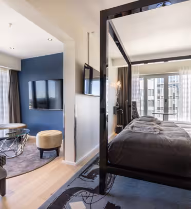 Lyxigt hotellrum med en himmelssäng, plyschkuddar och en mysig inredning. En sittgrupp inkluderar en modern stol, en ottoman, ett soffbord i glas och en stor TV monterad på en blå vägg. Fönster från golv till tak erbjuder utsikt över staden.