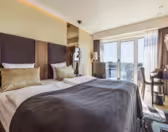 Modernt hotellrum med en stor säng, guld- och vita kuddar och en grå filt. Ett fönster erbjuder utsikt över staden, och det finns ett litet skrivbord med en stol till höger. Varm belysning och neutrala toner skapar en mysig atmosfär.