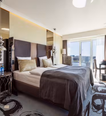 Modernt hotellrum med en stor säng, grå sängkläder och guldkuddar. Sänggavel med spegel, snygga nattduksbord och trägolv. Ett skrivbord och en stol finns nära stora fönster med skira gardiner, med utsikt över stadsbilden. Inredningen inkluderar hängande lampor och abstrakt matta.