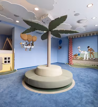 Lekrum för barn med blått och grönt färgschema, med en central tecknad palmträd, moln i taket och väggdekorationer av animerade karaktärer. Det finns ett litet hus i hörnet och röda gardiner med vita prickar.