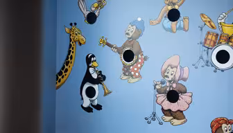 En lekfull väggmålning med tecknade djur som spelar instrument. En giraff med en trumpet, en pingvin med en tuba, en björn i en blå hatt med en gitarr och en kanin i en rosa klänning med en flöjt är avbildade. Det finns fem svarta cirkulära hål i väggmålningen.