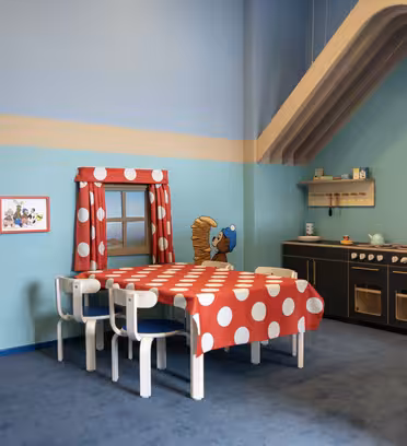 Lekrum för barn med blått och grönt färgschema, med ett köksset, en röd duk med vita prickar på ett bord med stolar och väggkonst av tecknade djur, inklusive en björn i en gungstol.