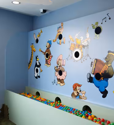 En färgglad lekplats har en väggmålning med tecknade djur som spelar musikinstrument. Under väggmålningen finns en bollgrop fylld med mångfärgade bollar. Rummet har ljusblå väggar och ett fönster till vänster.