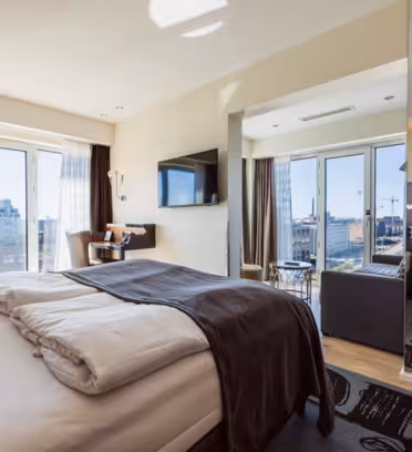 Modernt hotellrum med stor säng, mörka kuddar och sängkläder, TV, stadsutsikt från stora fönster, litet bord med stolar och en spegelvägg som speglar rummet. Ljusa trägolv och neutrala väggar skapar en mysig atmosfär.