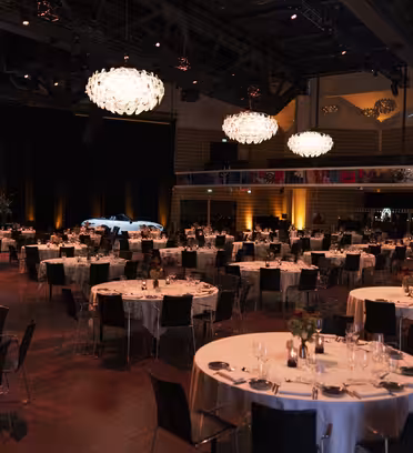 En stor festsal för ett event med runda bord täckta av vita dukar, var och en prydd med glas och bestick. Eleganta ljuskronor hänger i taket. En skärm visar "Van Mossel" och ett delvis synligt fordon står på scenen.