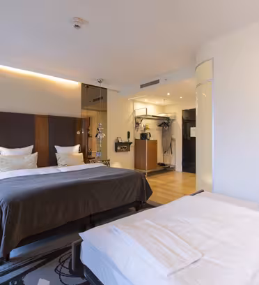 Ett modernt hotellrum med två snyggt bäddade sängar med vita sängkläder och bruna sängkläder. Rummet har trägolv, varm belysning och en väggmonterad TV. En glasvägg och en spegelvägg ger elegans och rymd till inredningen.
