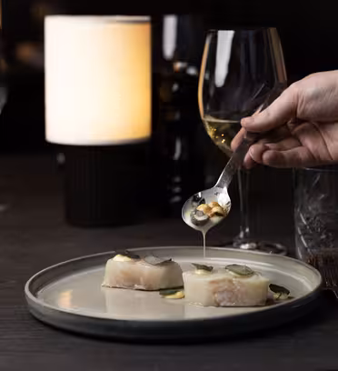 En hand som håller i en sked ringlar sås över två fiskbitar garnerade med skivor på en tallrik. Vinglas och en självlysande lampa skapar en elegant matsal på ett mörkt bord.