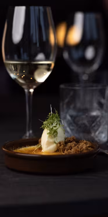 En matscen med svag belysning med en efterrätt på en tallrik garnerad med örter, ett glas vitt vin och ett glas vatten med is. En rullad servett med bestick ligger bredvid dem på ett mörkt träbord.