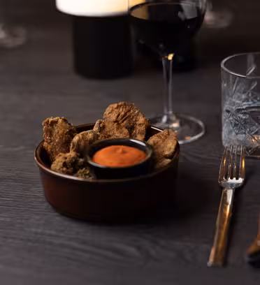 Ett träbord med en skål med stekt svamp, ett litet fat med röd sås, ett glas rött vin och ett glas vatten. En uppsättning gaffel och kniv finns bredvid skålen. Bakgrunden är svagt upplyst.