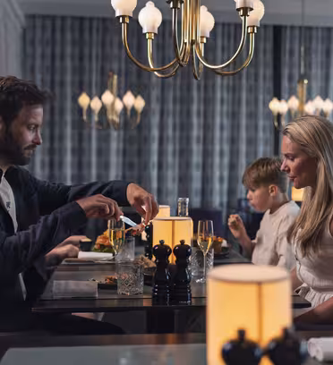 En familjemiddag på en restaurang. En man serverar sig själv från ett fat och sitter mitt emot en kvinna. Ett barn sitter bredvid kvinnan, fokuserat på sin måltid. Bordet har eleganta inredningar och atmosfären är varm med mjuk belysning.