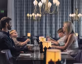 En familj på fyra sitter runt ett svagt upplyst restaurangbord; två vuxna och två barn möter varandra. Inramningen är elegant med moderna ljuskronor och varma lampor på bordet. Barnen verkar underhållas när de äter.
