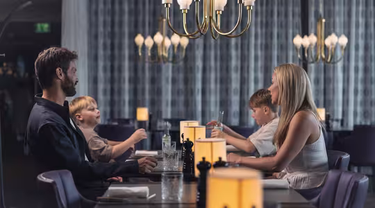 En familj på fyra sitter runt ett svagt upplyst restaurangbord; två vuxna och två barn möter varandra. Inramningen är elegant med moderna ljuskronor och varma lampor på bordet. Barnen verkar underhållas när de äter.