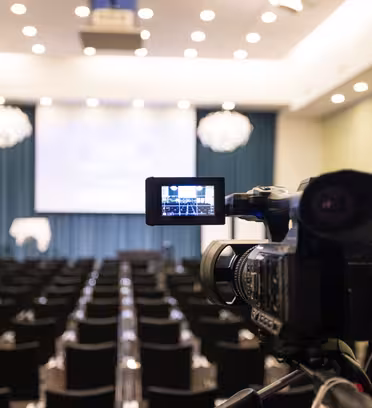 En videokamera är uppställd på baksidan av ett tomt konferensrum, vänd mot en scen med stor skärm. Rader av svarta stolar fyller rummet, och taket är prydt med ljuskronor. Rummet är mjukt upplyst, vilket tyder på beredskap för ett evenemang.