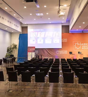 Ett konferensrum med rader av tomma stolar vetter mot en scen med ett podium och storbildsskärmar. På banners stod det "Rundabordssamtal för marknadsföringsinnovation." Skärmarna visar presentationsbilder med fyra högtalares huvudbilder. Kameror är inställda för inspelning.