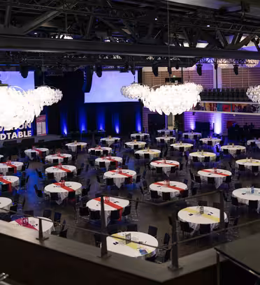 En stor konferenssal med runda bord dukade för ett event. Varje bord har en vit duk med färgade löpare. I taket hänger två stora skärmar med blå belysning och ljuskronor. "Innovation Roundtable" syns på scenen.