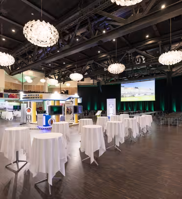 Vidvinkelvy av ett rymligt konferensrum med eleganta runda bord draperade i vita dukar. Stora ljuskronor hänger i taket och en stor skärm på scenen visar en presentation. Deltagarna är synliga i informell klädsel.