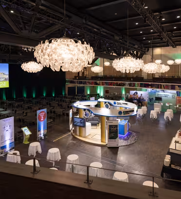 En rymlig konferenssal med eleganta ljuskronor är värd för ett evenemang. Montrar sätts upp med displayer och reklammaterial. Det finns bord med vita dukar och en stor skärm visar "Välkommen 2024." Stämningen är formell och inbjudande.