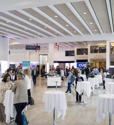 En rymlig kongresscenterlobby med människor som umgås och står runt höga, runda bord klädda i vita dukar. Olika bås och skyltar syns och det finns grönska i förgrunden.
