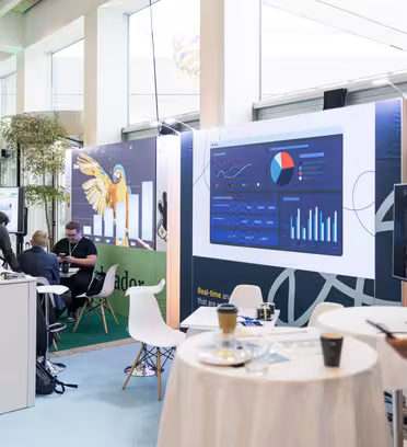 Ett modernt konferensområde med stora digitala skärmar som visar grafer och diagram. Människor interagerar vid minimalistiska bord med stolar. En man i förgrunden håller en mikrofon och papper. Naturligt ljus filtreras genom stora fönster.