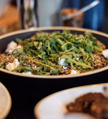 En sallad i en svart skål med linser, getost och ruccola. Runt den står andra rätter, något oskarpa, på en mörk bordsskiva. Bakgrunden visar suddiga köksartiklar.