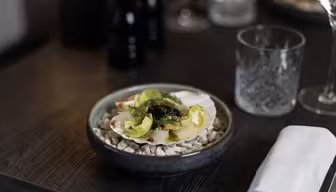 En skål med pilgrimsmusslor med grön garnering presenteras på en bädd av stenar i en mörk skål. Den placeras på ett träbord tillsammans med ett glas, en servett och suddiga bakgrundselement, inklusive en ljus- och pepparkvarn.