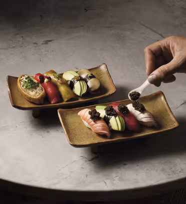 En hand lägger försiktigt kaviar på en bit sushi på ett rektangulärt keramiskt fat. Båda skålarna består av olika färgglada sushibitar arrangerade på en strukturerad stenyta.
