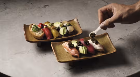 En hand lägger försiktigt kaviar på en bit sushi på ett rektangulärt keramiskt fat. Båda skålarna består av olika färgglada sushibitar arrangerade på en strukturerad stenyta.