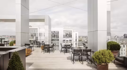 Takterrass med modern arkitektur med höga vita pelare och glasräcken. Området har svarta bord och stolar ordnade prydligt, krukväxter för dekoration och utsikt över stadsbyggnader i bakgrunden under en molnig himmel.