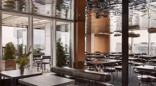 Modern matplats inomhus med svarta bord och stolar, med reflekterande tak. Stora fönster visar upp en utomhusterrass med grönska och utsikt över staden. Inredningen inkluderar läderstolar och moderna belysningsarmaturer.