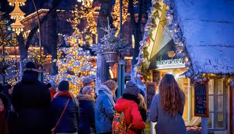 En festlig utomhusjulmarknadsscen med människor samlade runt ett dekorerat stånd. Varma lampor och hängande lyktor lyser upp området. Ett snötäckt tak förhöjer den mysiga vinterstämningen och träden pryds av glödande ljusslingor.
