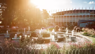Bilden visar en solbelyst trädgård med en cirkulär fontän omgiven av blommor. I bakgrunden finns en byggnad med en rund fasad och rader av flaggor ovanpå. Scenen är levande och stämningsfull.