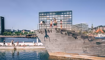 Människor är samlade på en träkonstruktion vid en strand, och några förbereder sig för att dyka i vattnet. Moderna byggnader och klar himmel finns i bakgrunden. Graffiti är synligt på strukturen.