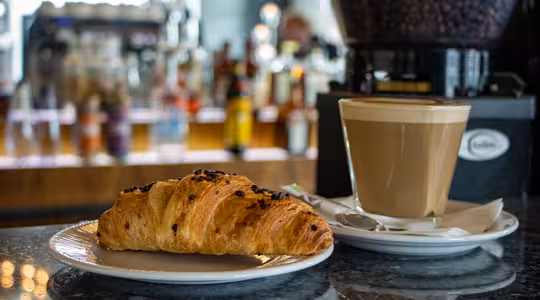 En flagnande croissant på en vit tallrik står bredvid ett glas skummande kaffe på en kafédisk. Bakom dem visar suddiga hyllor olika drycker på flaska. Den varma belysningen skapar en mysig atmosfär.
