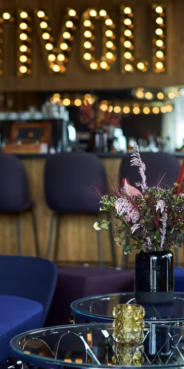 En elegant lounge med blå och gula stolar, glasbord och en blommig mittpunkt. Bakgrunden har en bar med lila pallar och en tältskylt som läser "TIVOLI" med upplysta bokstäver.