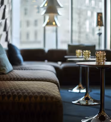 En elegant loungedel med mönstrade soffor, blå kuddar och runda metalliska bord, var och en toppad med dekorativa föremål. Utrymmet är mjukt upplyst av moderna taklampor, med ett stort fönster som visar en suddig stadsbild utanför.